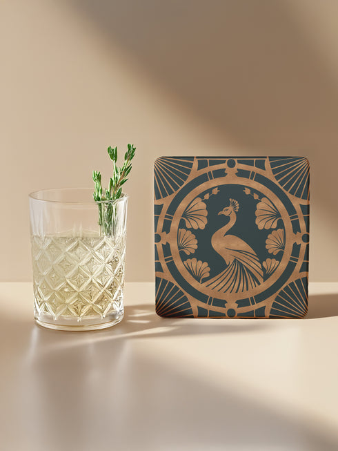 Square ceramic coaster featuring an Art Deco copper peacock design centered in a medallion, surrounded by fan patterns and shell motifs on a dark forest green background, with cork backing.